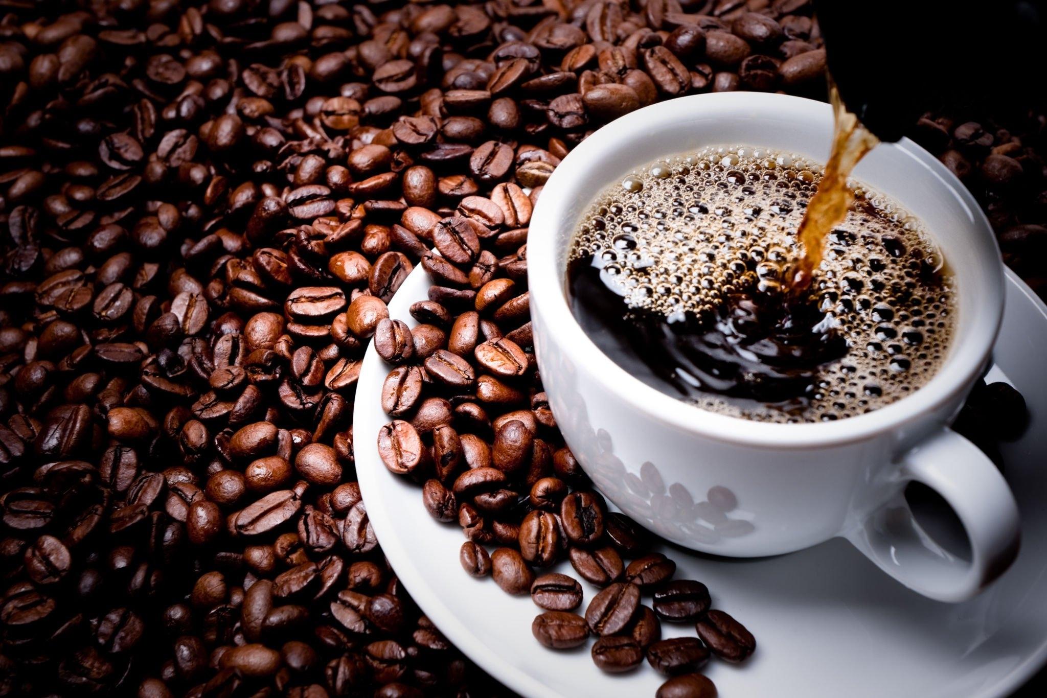 Hot coffee being poured into a white cup surrounded by roasted coffee beans